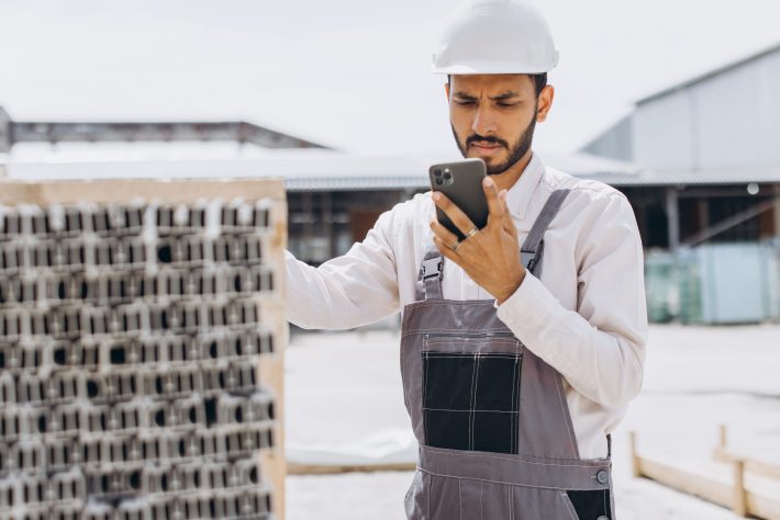 Construction operative in hard hat using smartphone to complete equipment fault reporting on a construction site