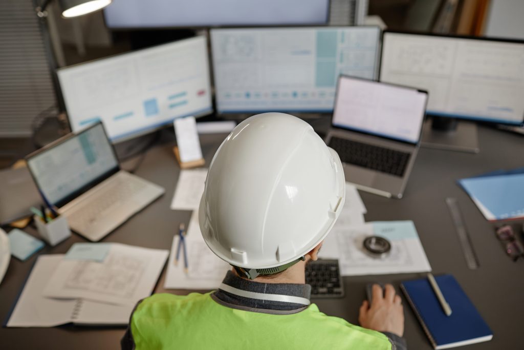 Fleet manager in a hi-vis vest and hard hat reviewing data on multiple monitors in a transport office