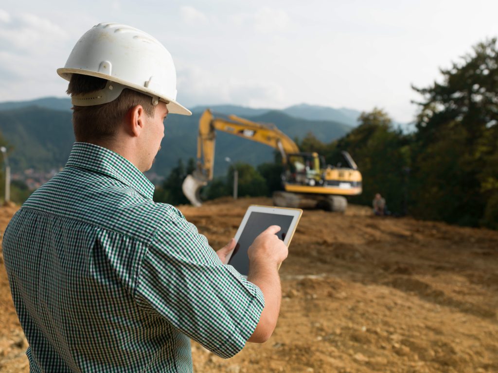 Construction site manager reviewing pre-use inspection software on a tablet with an excavator working in the background