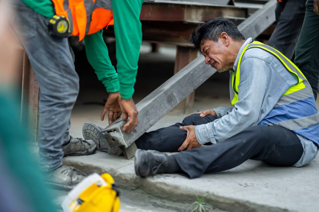 Construction worker sitting on the ground holding his leg after a steel beam fell, colleagues helping