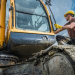 Worker in a hard hat and gloves opening the cab door of an excavator